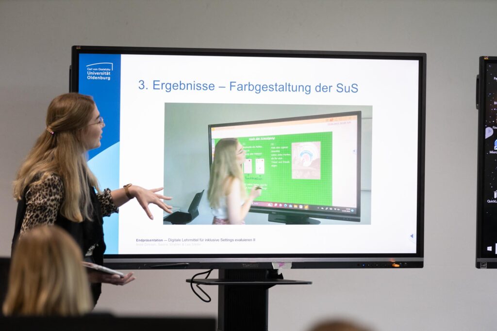 Eine Frau steht vor einem Bildschirm, auf dem wiederum eine Frau, die vor einem Bildschirm steht, abgebildet ist.